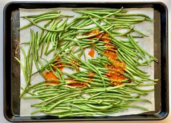 pour harissa sauce over green beans on a parchment lined baking sheet
